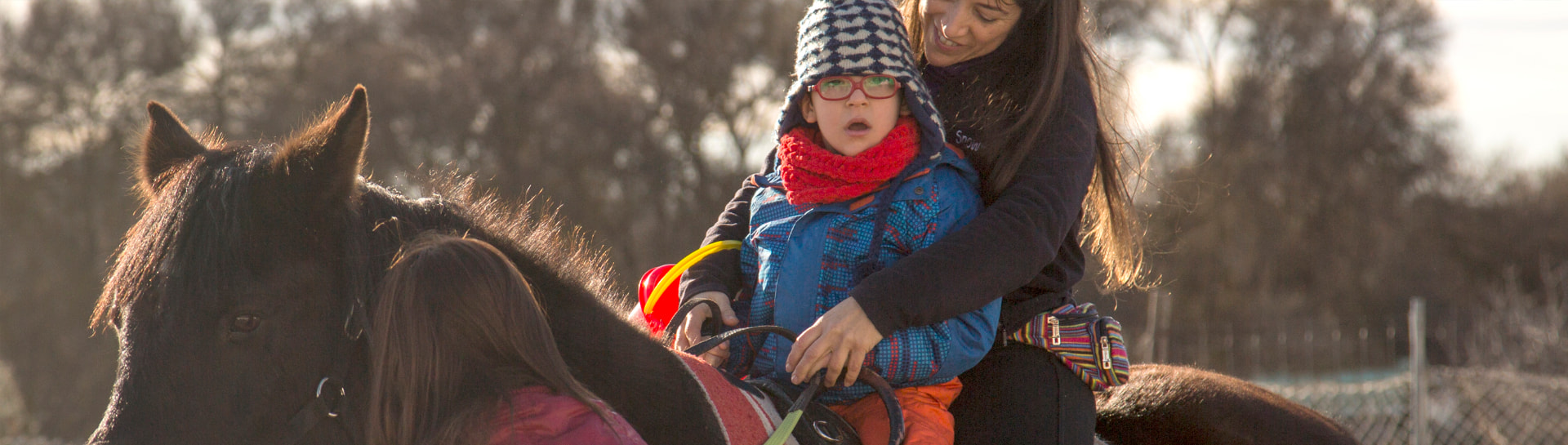 Niño con gorro y gafas montando a caballo acompañado por una terapeuta y una niña que lleva al poni
