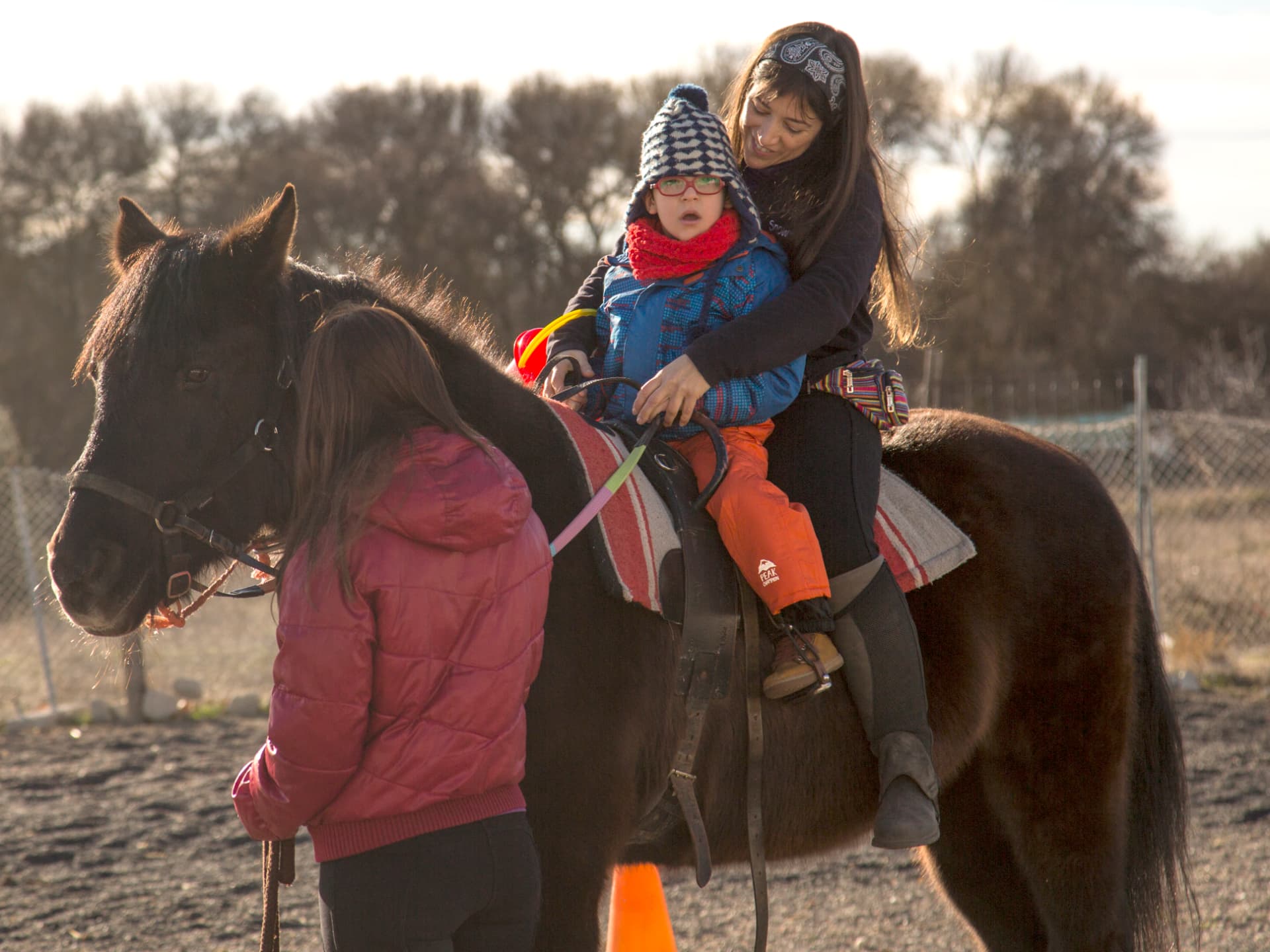 Niño con gorro y gafas montando a caballo acompañado por una terapeuta y una niña que lleva al poni