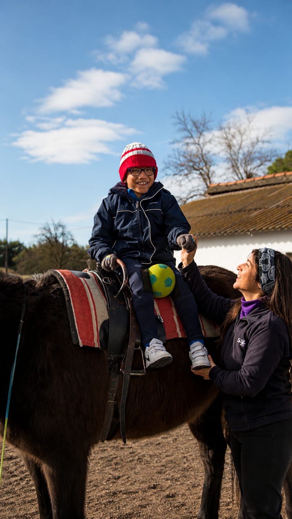 Niño sonriente con gorro y gafas montado a caballo acompañado por una terapeuta